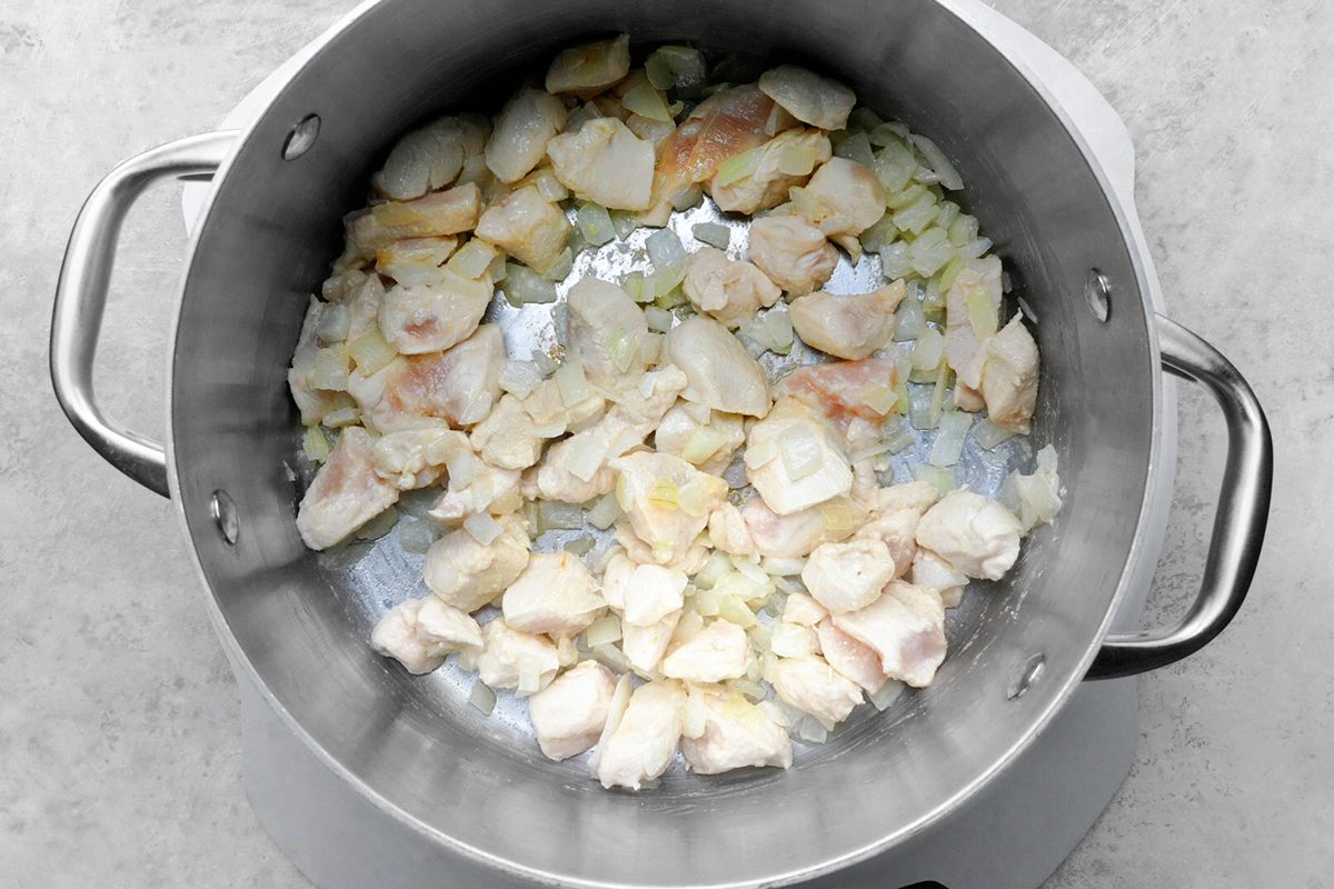 A steel pot on a stove containing chopped onions and diced chicken pieces being sautéed. The ingredients are lightly browned, creating a savory base. The pot has sturdy handles on each side.