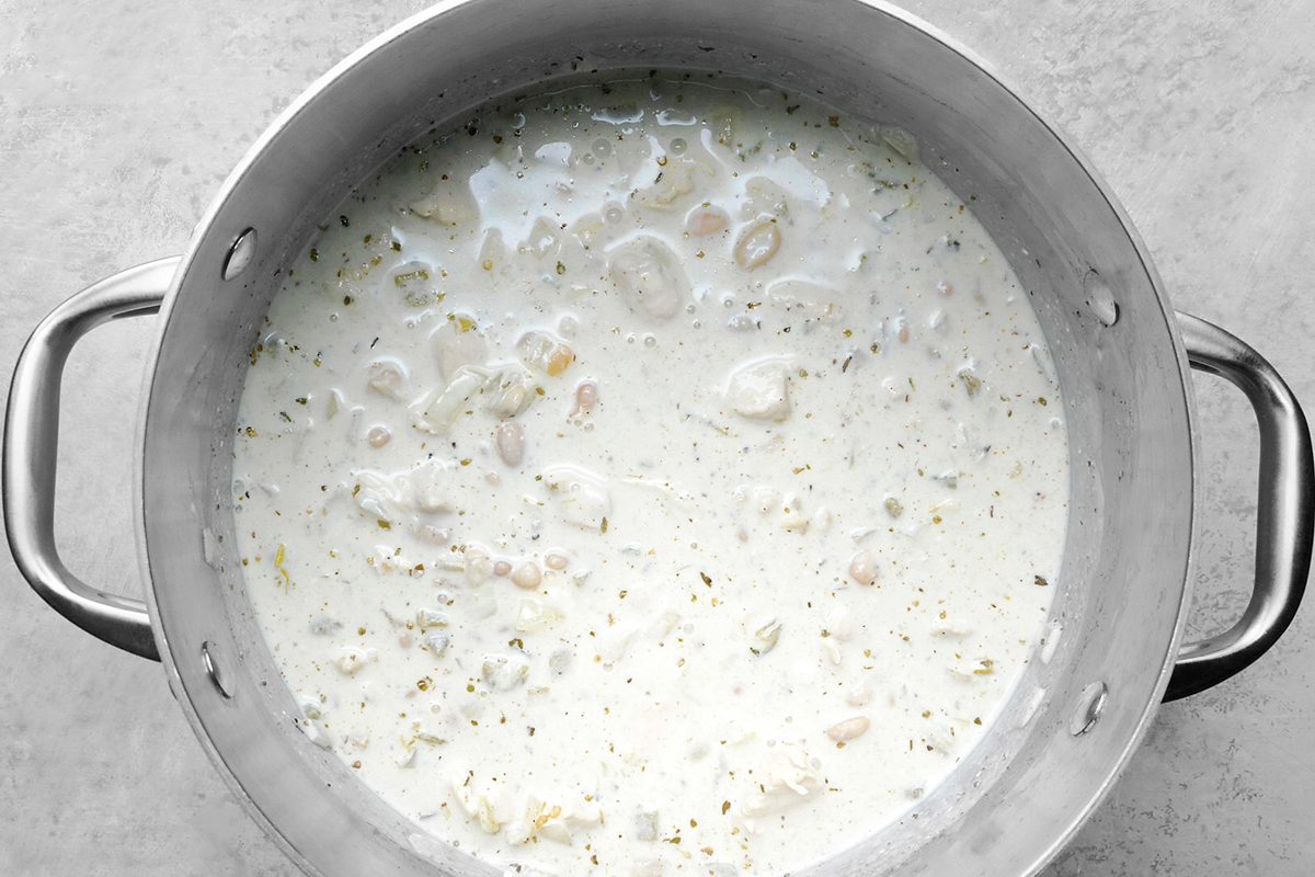 A large metal pot filled with a creamy white soup containing pieces of chicken, beans, and herbs, placed on a light gray surface.