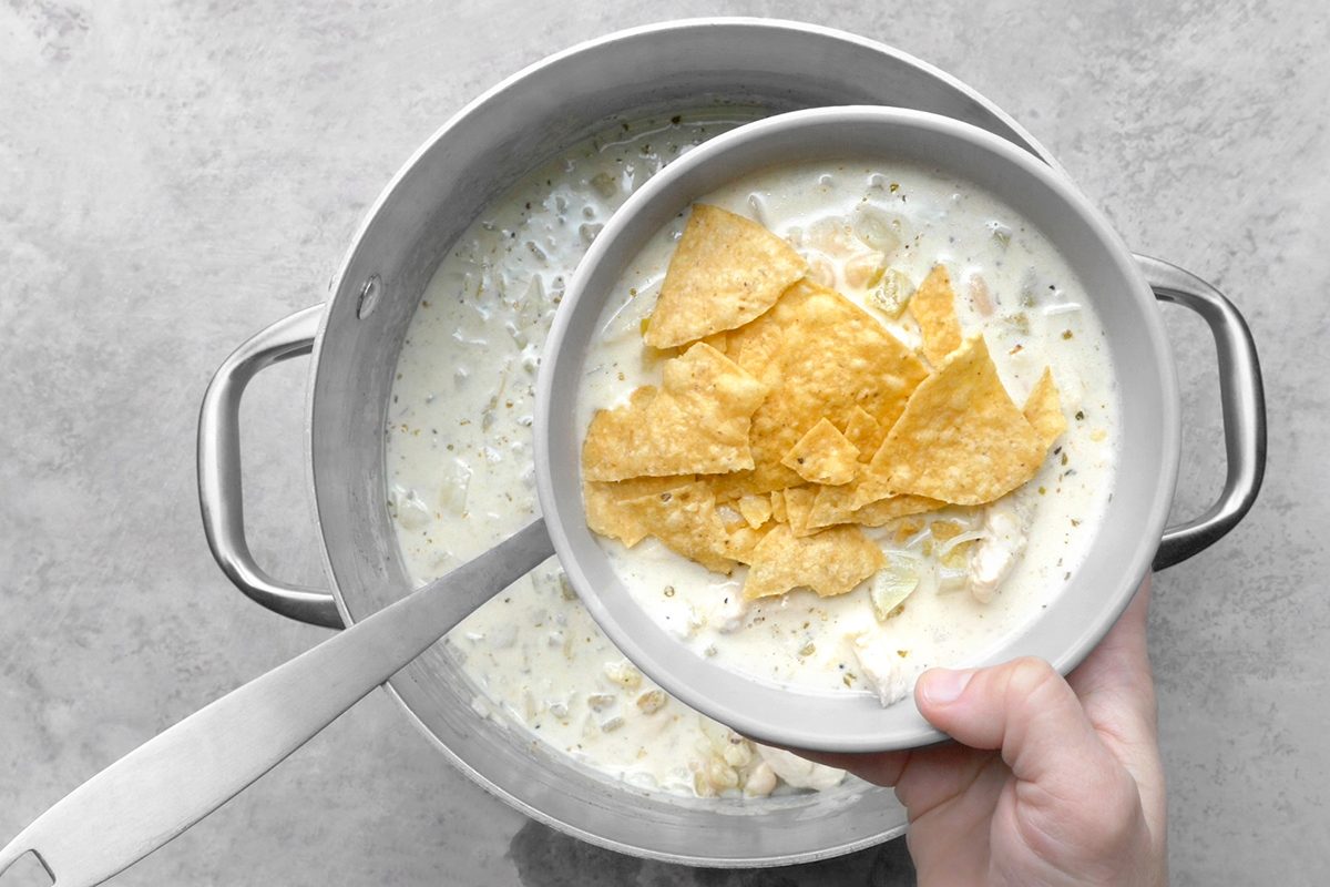 A hand holding a bowl of creamy soup topped with broken tortilla chips. The bowl is being held over a pot filled with more of the same soup, and a ladle rests on the edge of the pot. The background is a light gray surface.