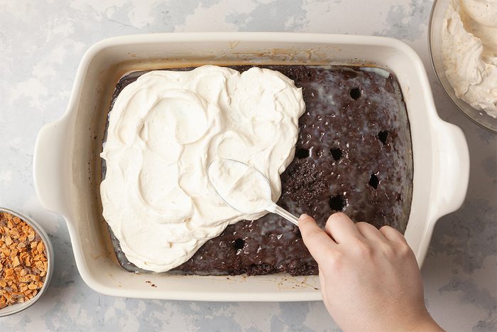 A hand spreads whipped cream over a chocolate poke cake in a rectangular dish. Holes in the cake are visible. A small bowl of toffee bits is on the left, and a bowl with more whipped cream is on the right. The background is a light textured surface.