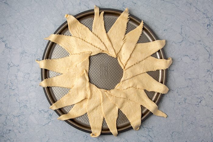 A circular baking tray with crescent roll dough arranged to form a ring, with the pointed ends facing outward. The light dough contrasts with the gray background surface.