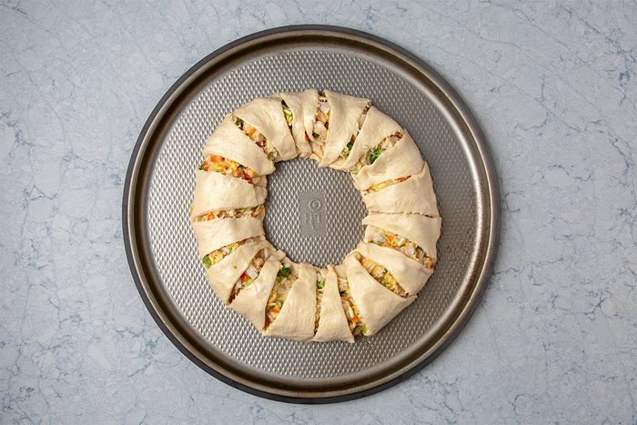 A round, unbaked, stuffed pastry wreath is on a circular baking sheet. The dough is folded over filling that includes vegetables and cheese, arranged in segments, ready for baking. The background is a light marbled surface.