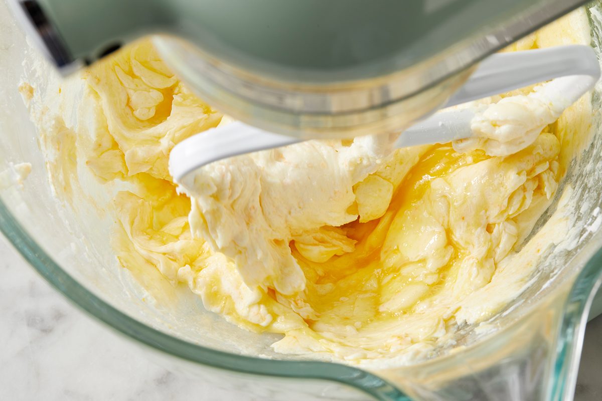 A close-up of a glass mixing bowl with creamy, pale yellow cake batter being mixed by a stand mixer. The batter appears smooth and whipped, with a paddle attachment partially visible. The bowl is set on a light marble surface.