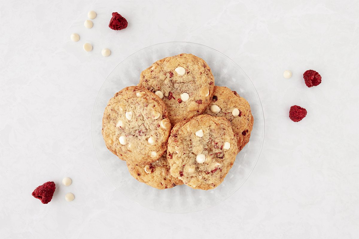 A plate of cookies with white chocolate chips, surrounded by scattered raspberries and white chocolate chips on a light surface.