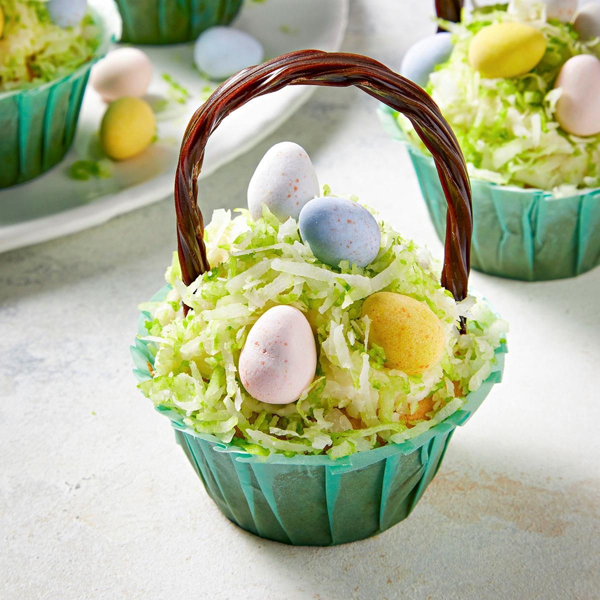 A cupcake decorated to look like an Easter basket, with green shredded coconut as grass, pastel-colored candy eggs on top, and a handle made from braided chocolate, in a teal wrapper. Other similar cupcakes are blurred in the background.
