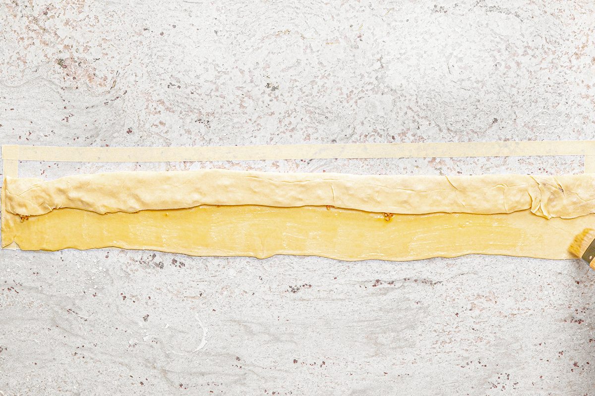 A strip of rolled-out dough is laid on a marble surface. It is being brushed with a light coating of egg wash or butter using a pastry brush. Another thin strip of dough is placed above the rolled dough.