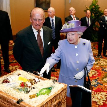 Queen Elizabeth II in a lavender dress points at a basket full of foods inside a banquet hall