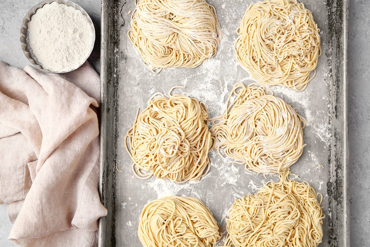 Five nests of raw noodles on a floured baking sheet. A bowl of flour and a pink cloth are placed on the left side. The noodles and sheet have a dusting of flour, giving a homemade feel. The background is a light, textured surface.