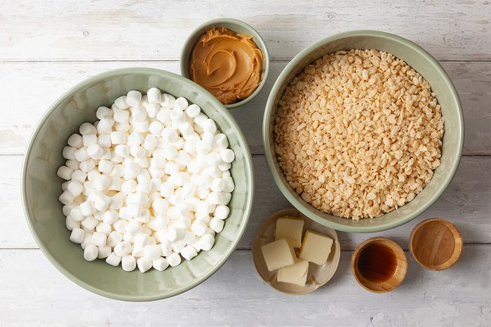Bowls on a wooden table contain ingredients for a sweet treat. Large bowl of mini marshmallows, another with crispy rice cereal, a small bowl of peanut butter, a few cubes of butter, and a small bowl with vanilla extract.