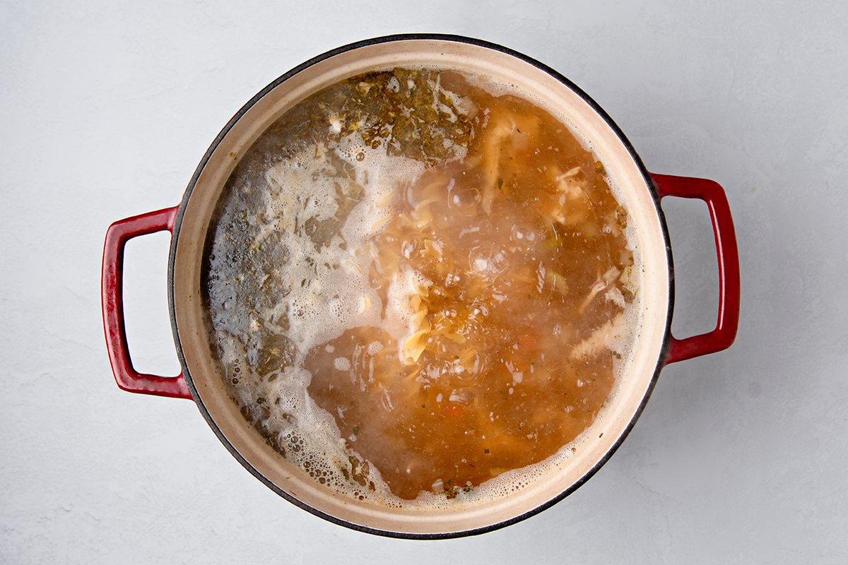 A red-handled pot filled with boiling broth and vegetables sits on a light gray surface. The liquid is bubbling, with ingredients like herbs and possibly pieces of chicken visible.
