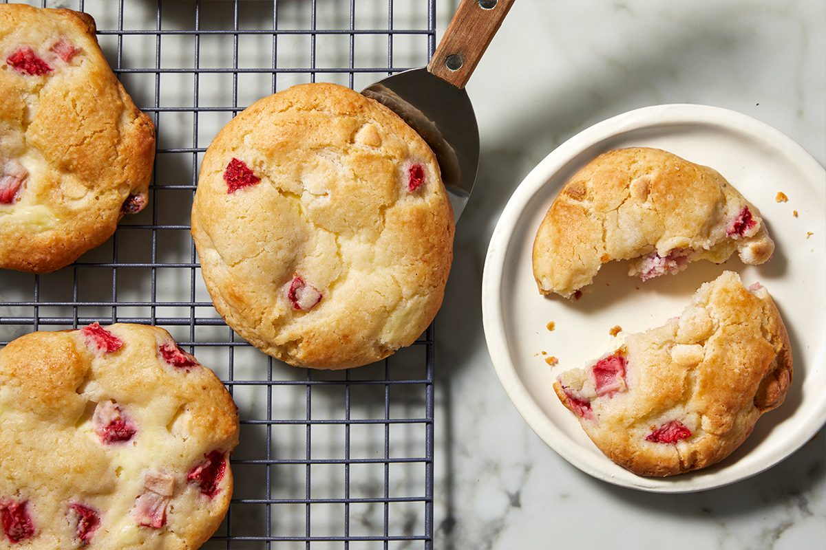 Overhead view of a spatula serving strawberry cheesecake cookies from a cooling mesh rack to a white plate where a broken cookie lies.