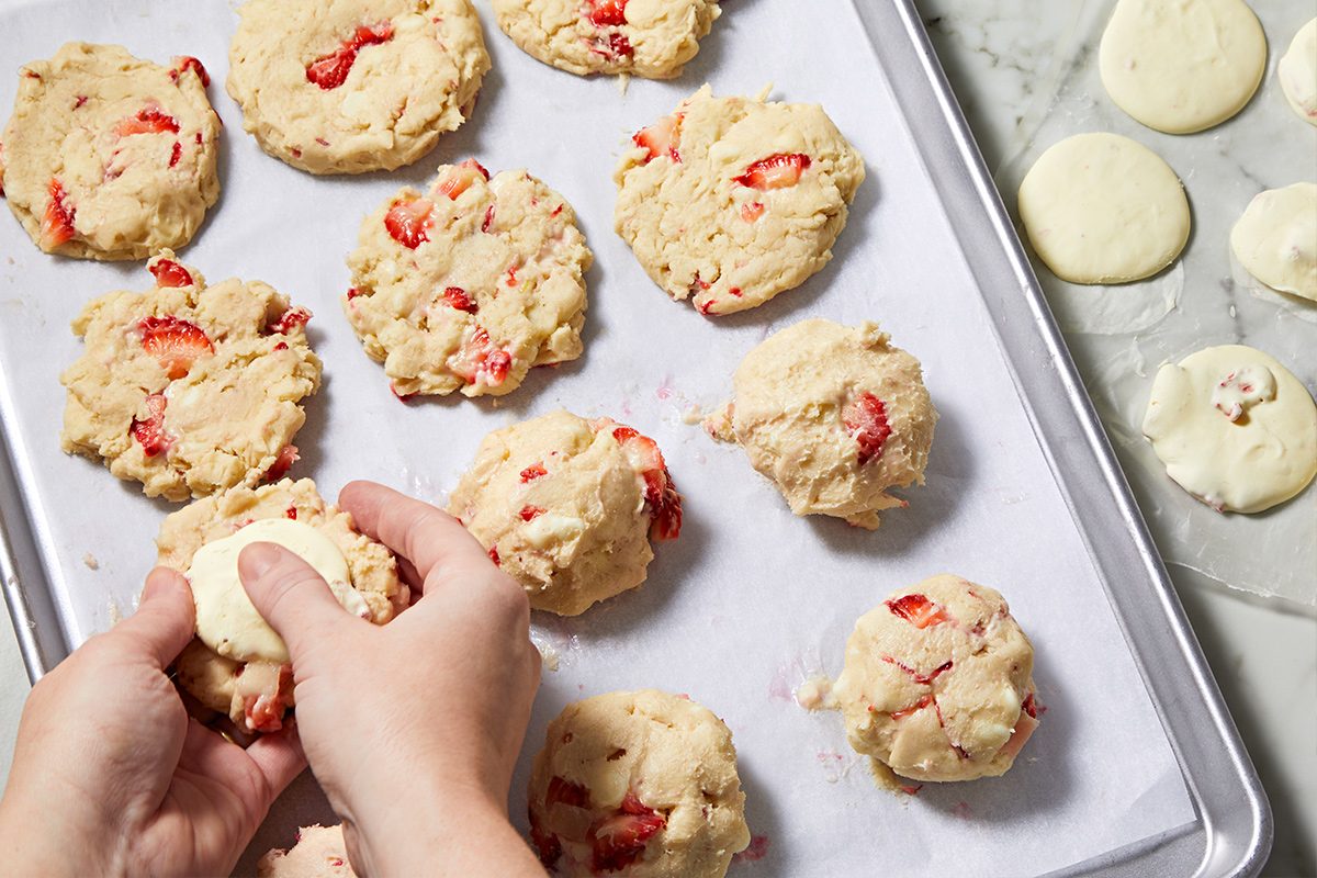 Overhead view of hands placing a ball of frozen cream cheese into the flattened cookie dough onto a parchment paper-lined baking sheet.